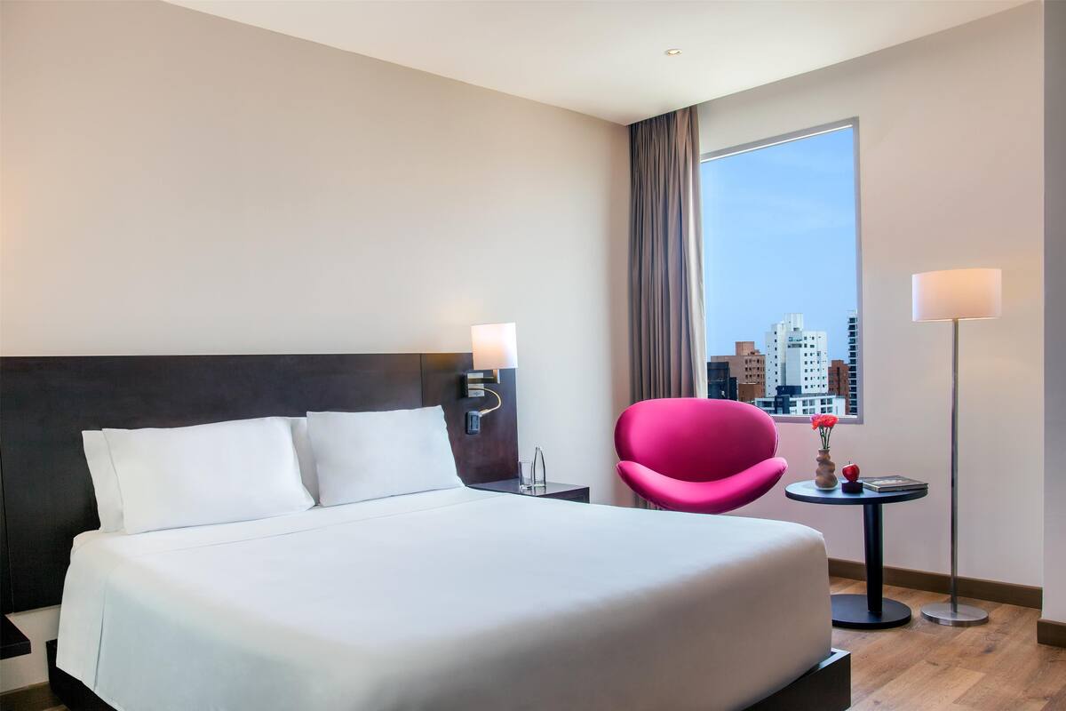 A hotel room with a large bed, a nightstand with a lamp, a small table with a vase and a book, a pink chair, and a view of the city through a window.