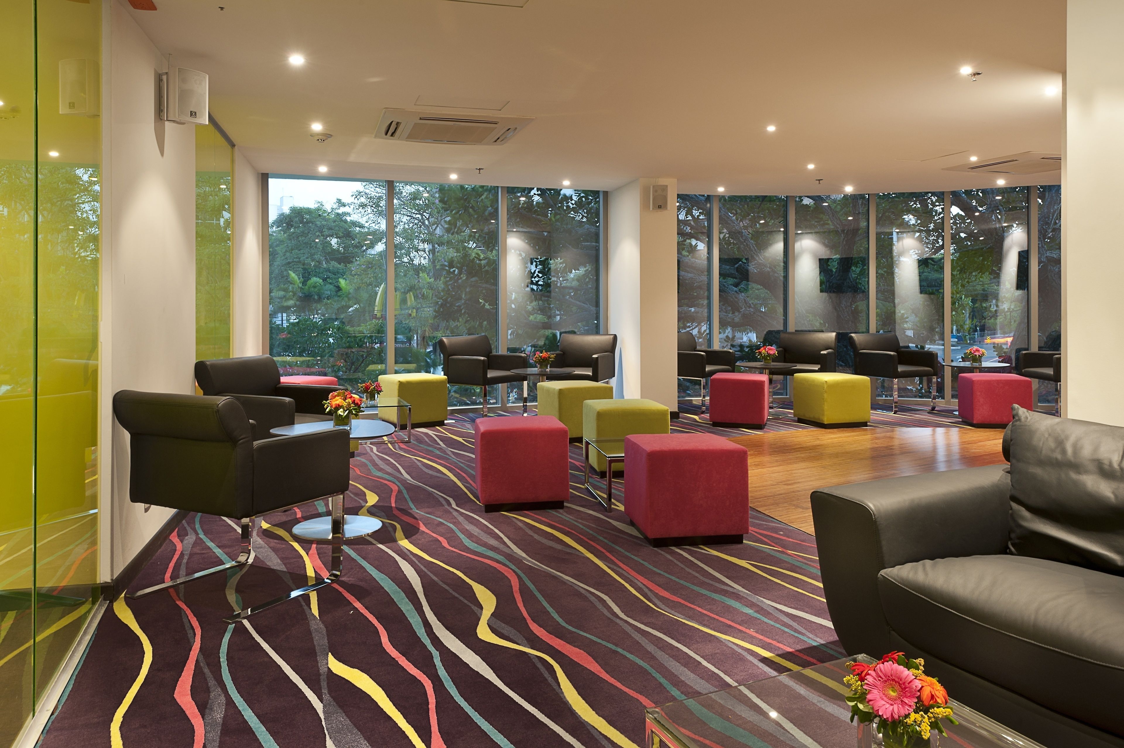 A modern lounge area with multiple seating options, large windows offering natural light and views of greenery, and a patterned carpet.