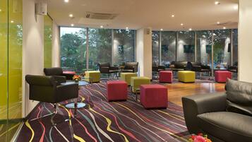 A modern lounge area with multiple seating options, large windows offering natural light and views of greenery, and a patterned carpet.