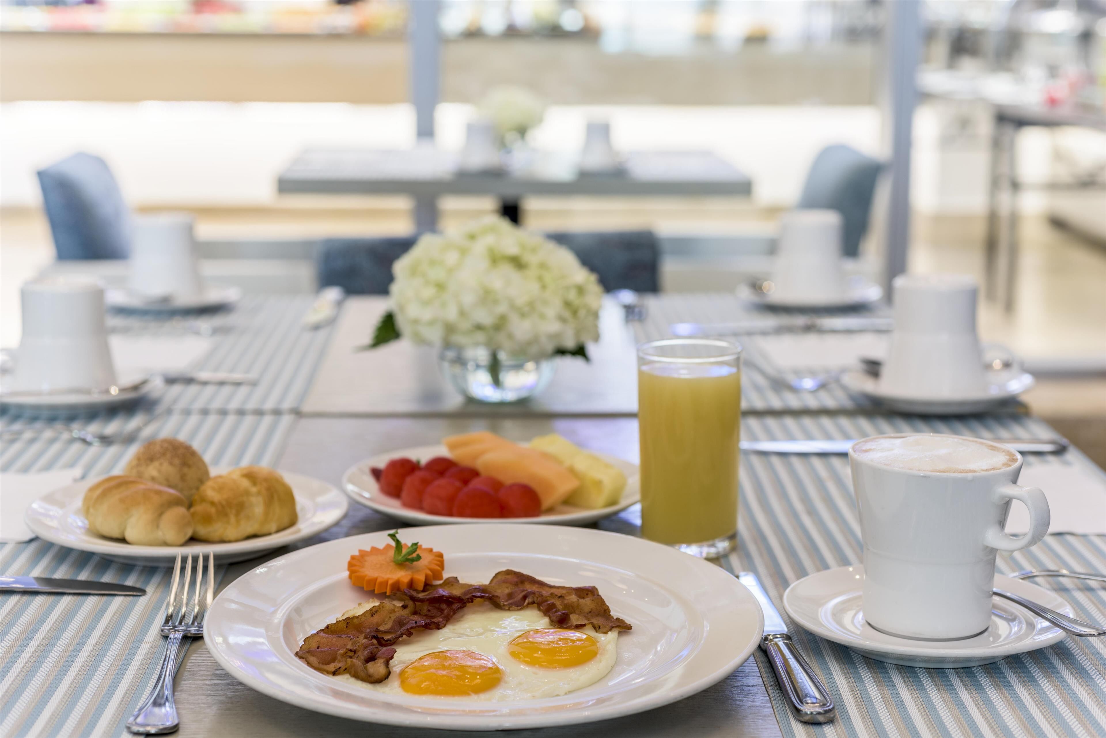Desayuno con cruasanes, huevos, tocino, fruta y café en una mesa.
