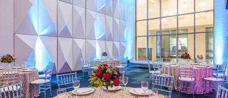 Un salón de banquetes con mesas redondas dispuestas para comer, sillas traslúcidas y ventanas amplias que dejan entrar luz natural.