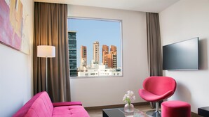 A modern living room with a pink sofa, a red chair, a glass coffee table, and a view of tall buildings through a large window. - NH Collection Barranquilla Smartsuites Royal (Barranquilla)