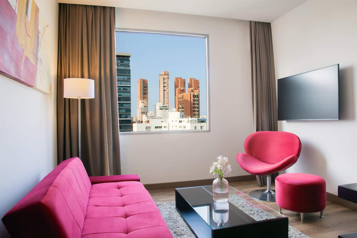 A modern living room with a pink sofa, a red chair, a glass coffee table, and a view of tall buildings through a large window.