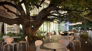 An outdoor seating area with round tables and white chairs, surrounded by a large tree and potted plants. - NH Collection Barranquilla Smartsuites Royal (Barranquilla)