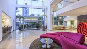 A spacious lobby with a modern design, featuring a large seating area with a pink sofa and a round table, large windows offering views of the city, and a glass railing on the upper level. - NH Collection Barranquilla Smartsuites Royal (Barranquilla)