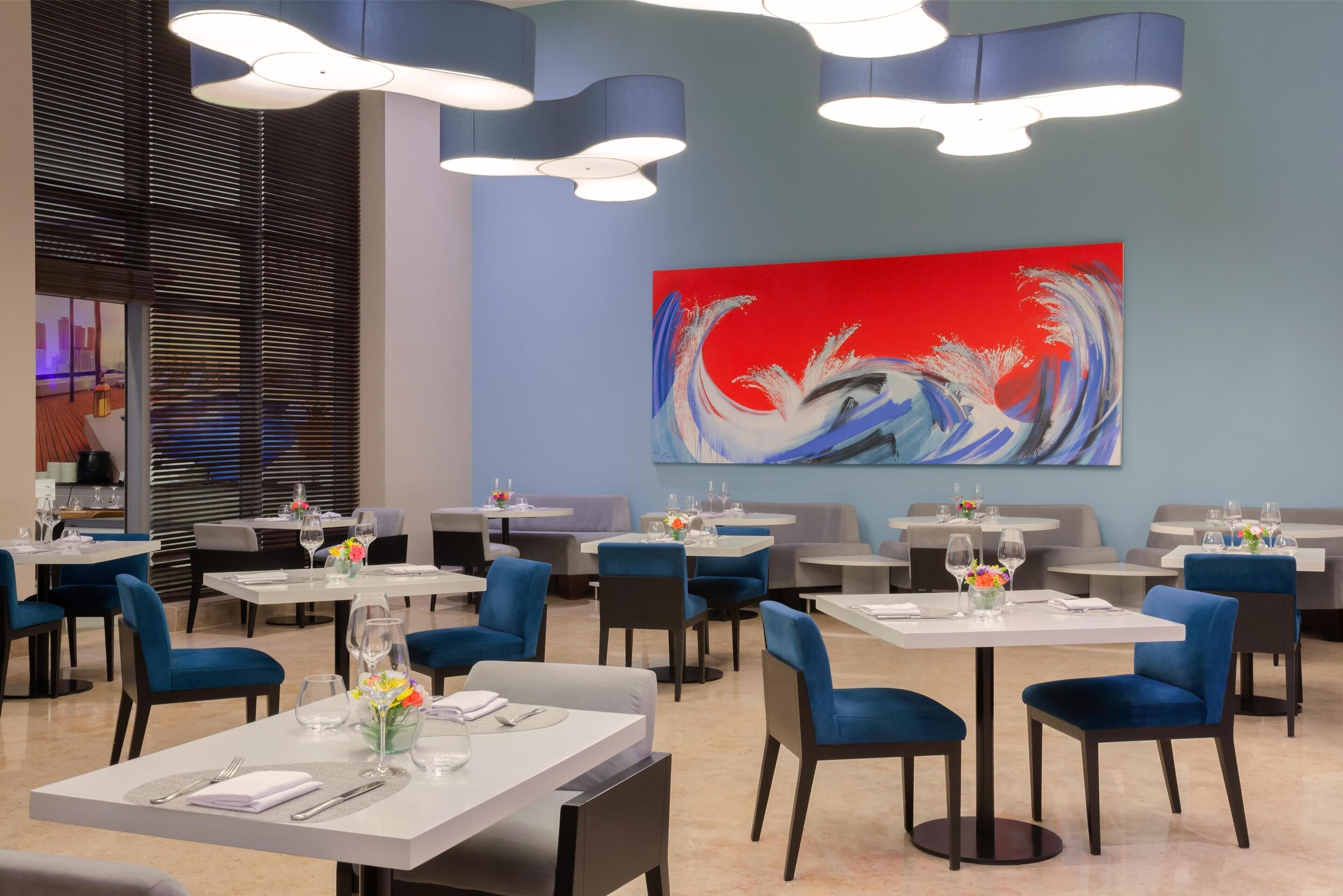 A modern restaurant interior with round tables, blue chairs, and a large abstract painting on the wall.