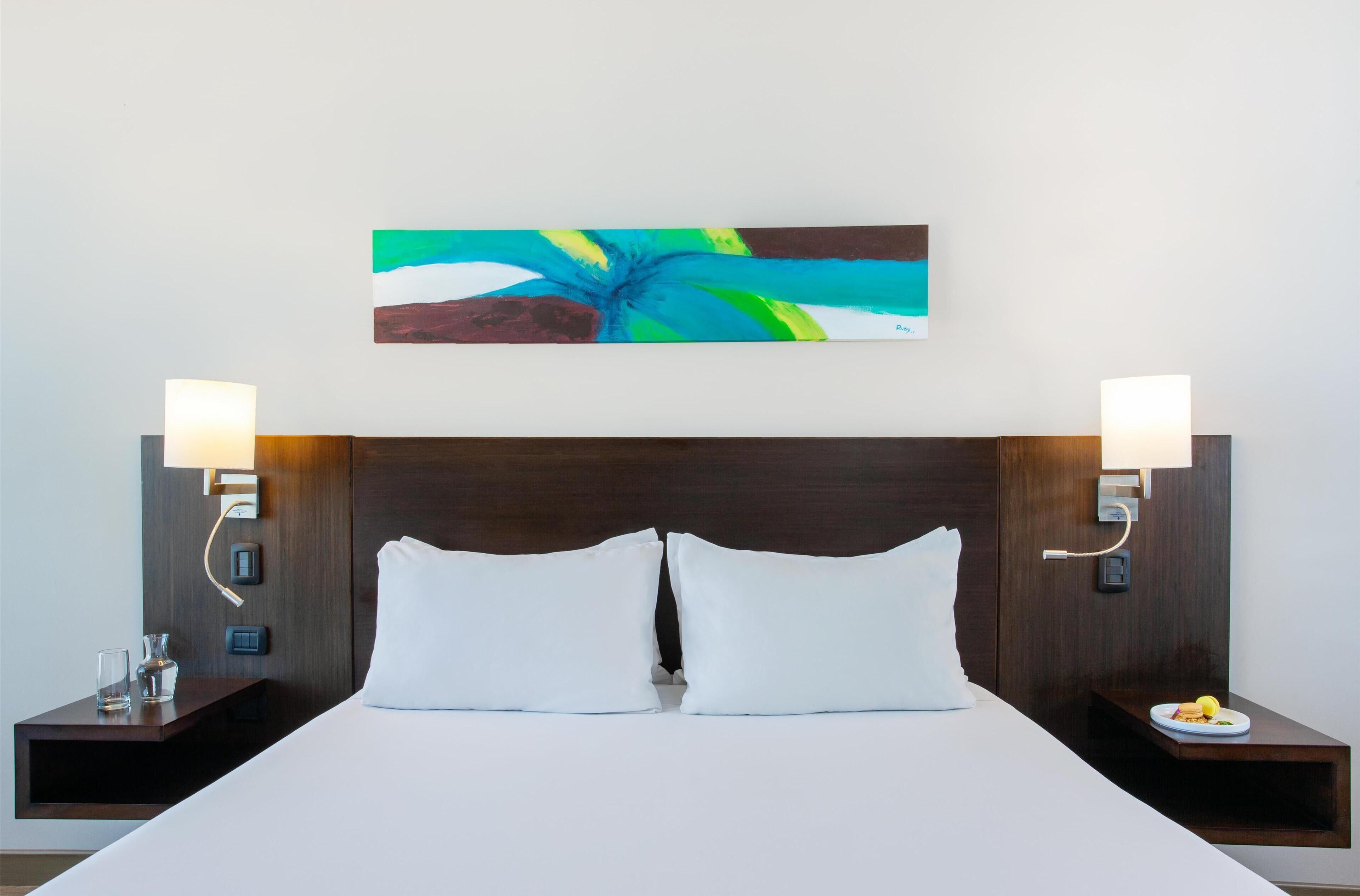 A neatly made bed with white linens, two bedside tables with lamps, and a colorful abstract painting on the wall.