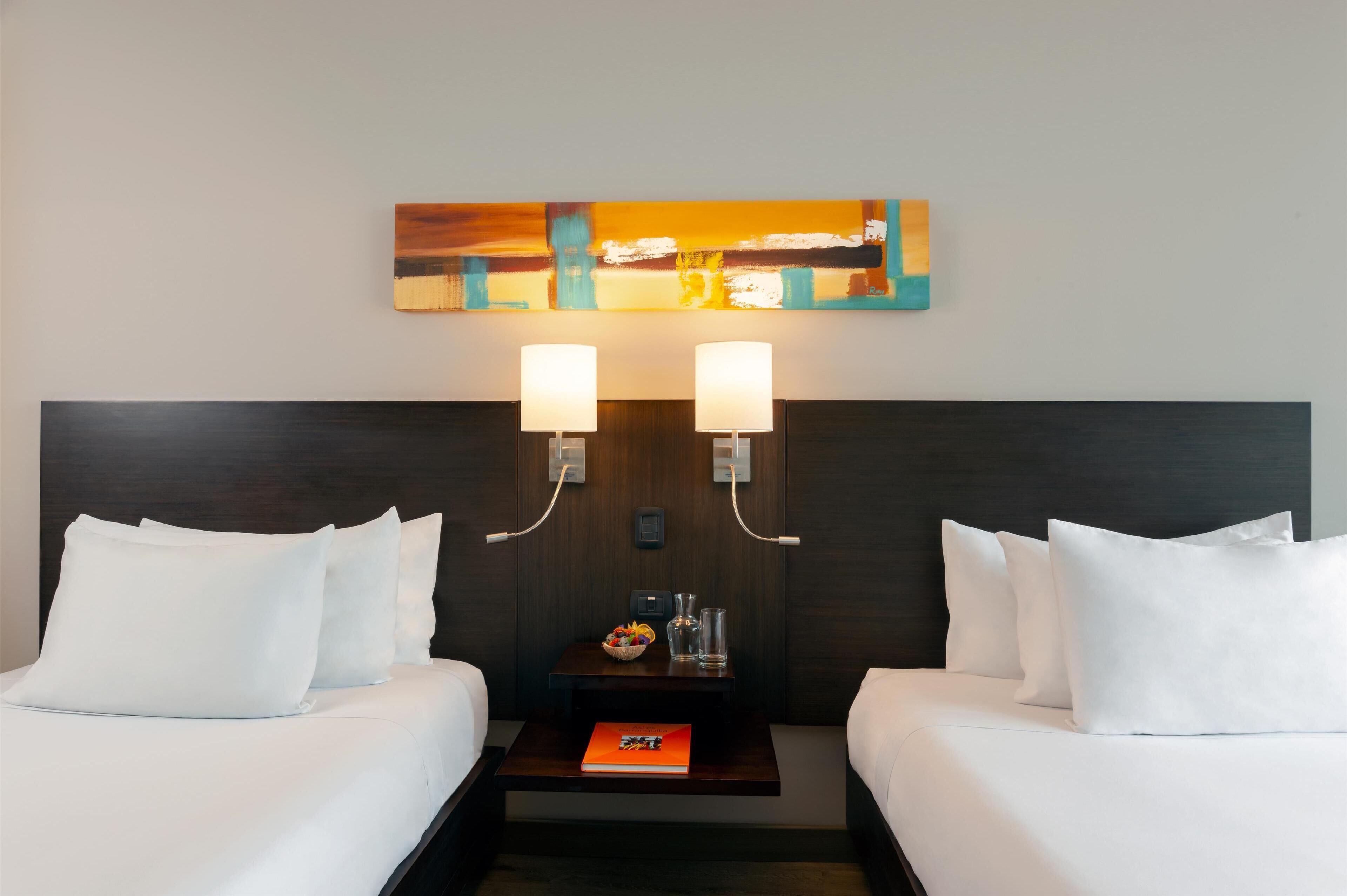 A hotel room with two beds, a nightstand, a painting, and two wall-mounted lamps.