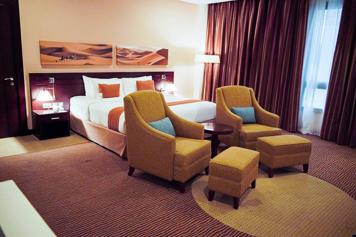 Habitación de hotel con una cama grande, dos sillones, un pequeño taburete, una mesita de noche con una lámpara y un cuadro de un paisaje desértico en la pared.