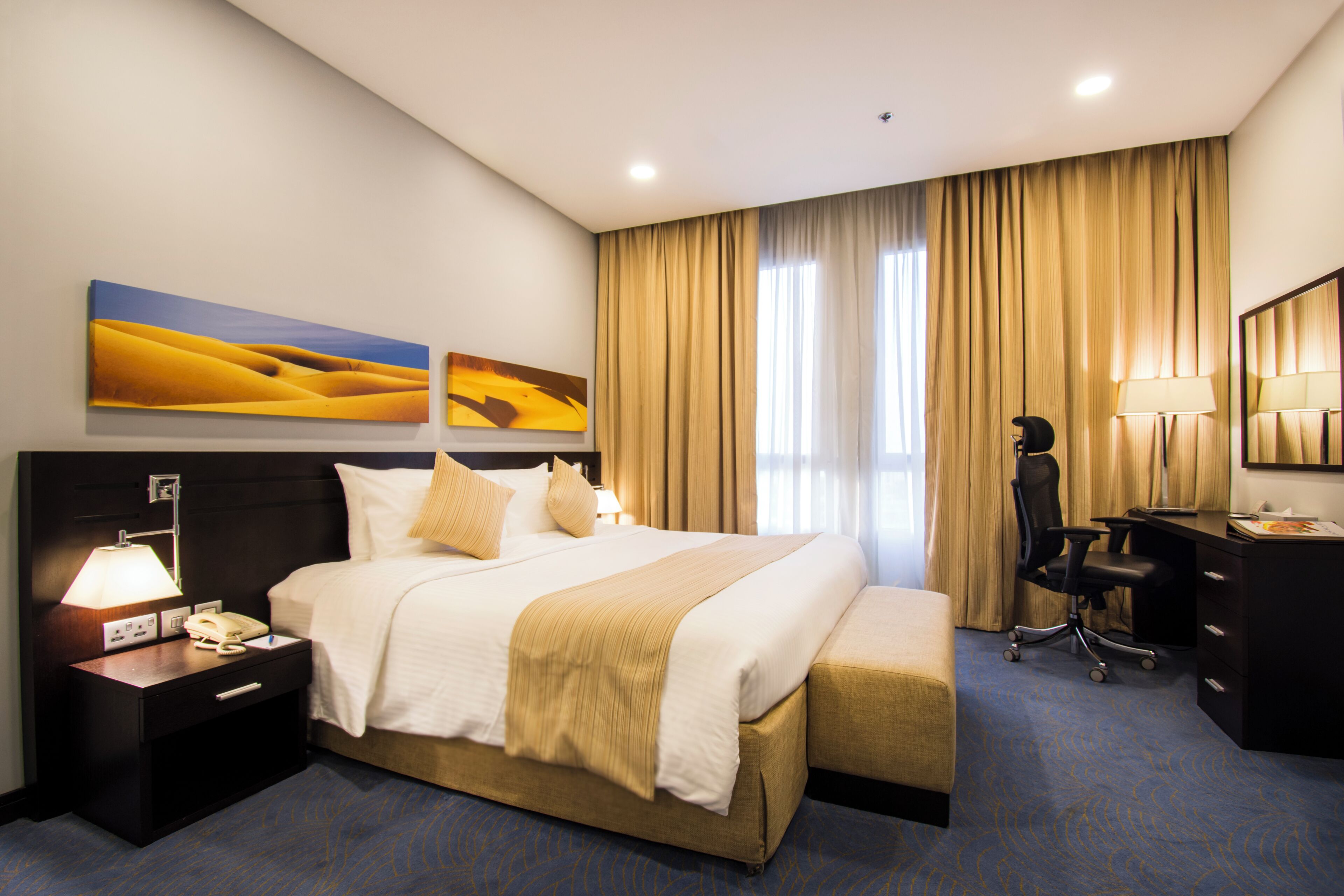A hotel room with a large bed, a desk, a chair, and two paintings on the wall.