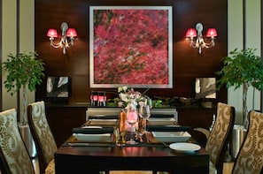 A dining area with a table set for two, flanked by patterned chairs, a large abstract painting on the wall, and wall-mounted lamps. - City Seasons Hotel Muscat (Muscat)