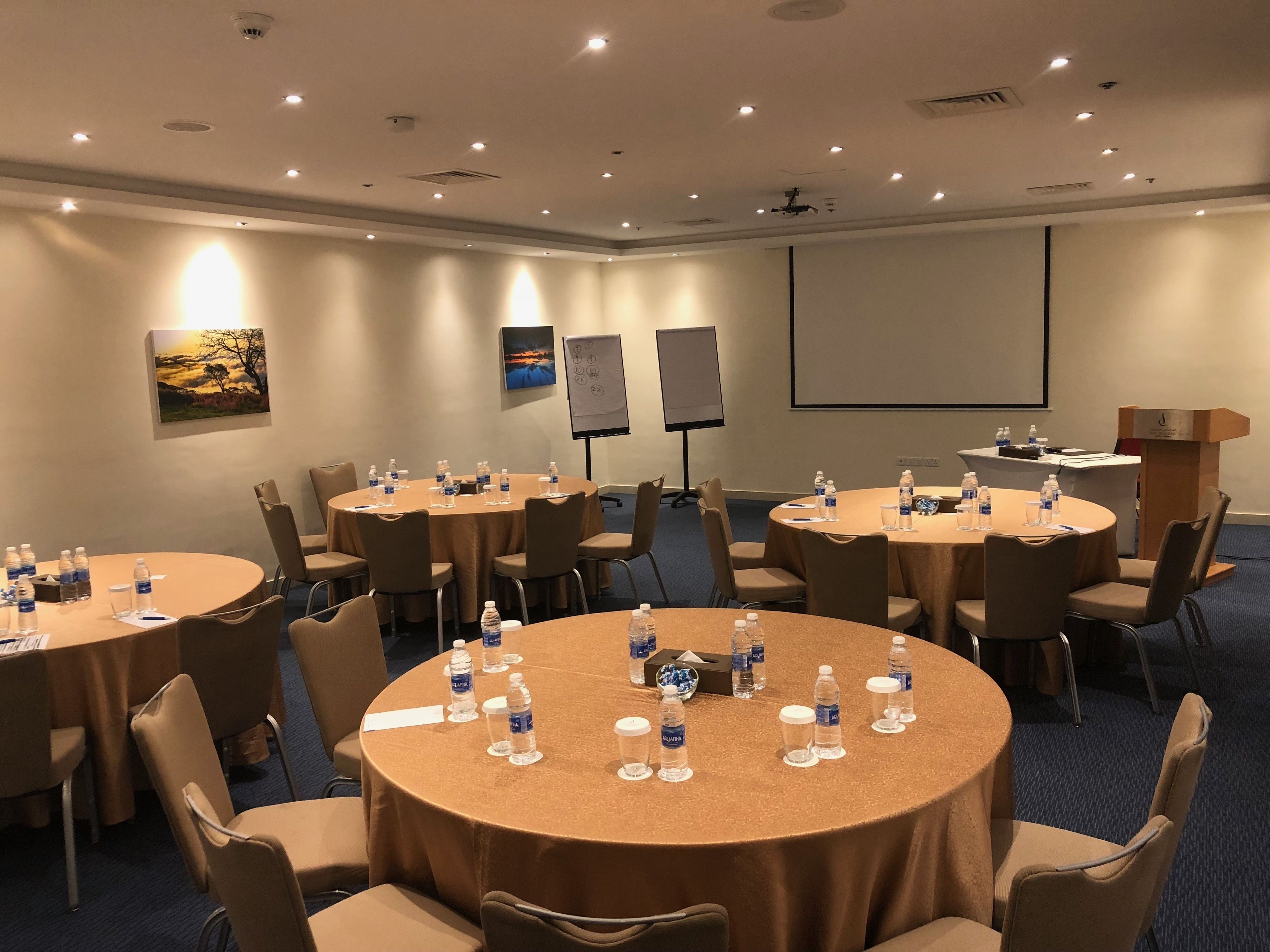 Une salle de conférence dotée de plusieurs tables rondes, chacune étant pourvue de bouteilles d’eau et de verres, et équipée d’un écran de projection et de panneaux de présentation.