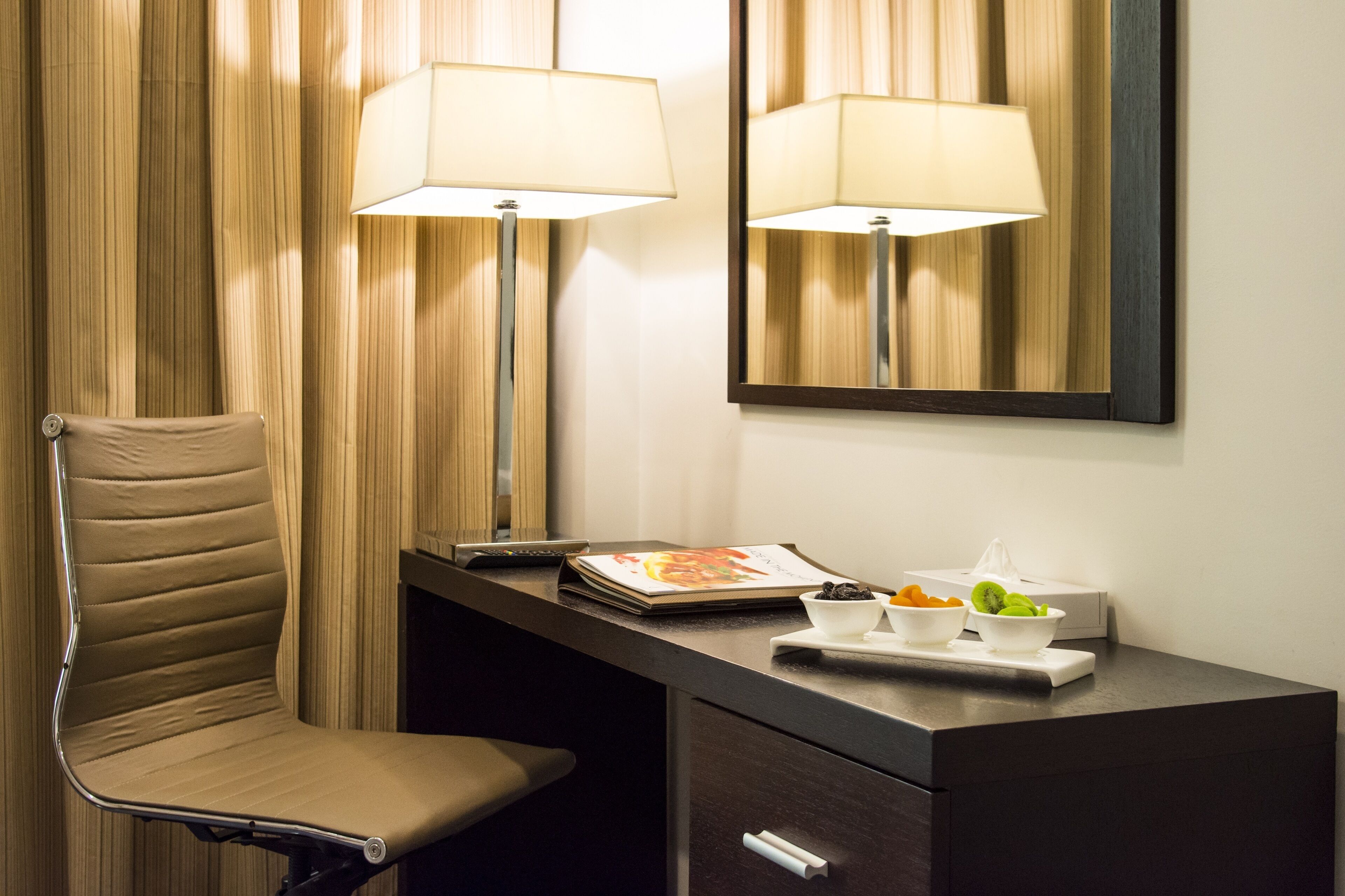 Habitación de hotel con una silla acolchada, un escritorio con lámpara, un espejo y una bandeja con fruta y un libro.