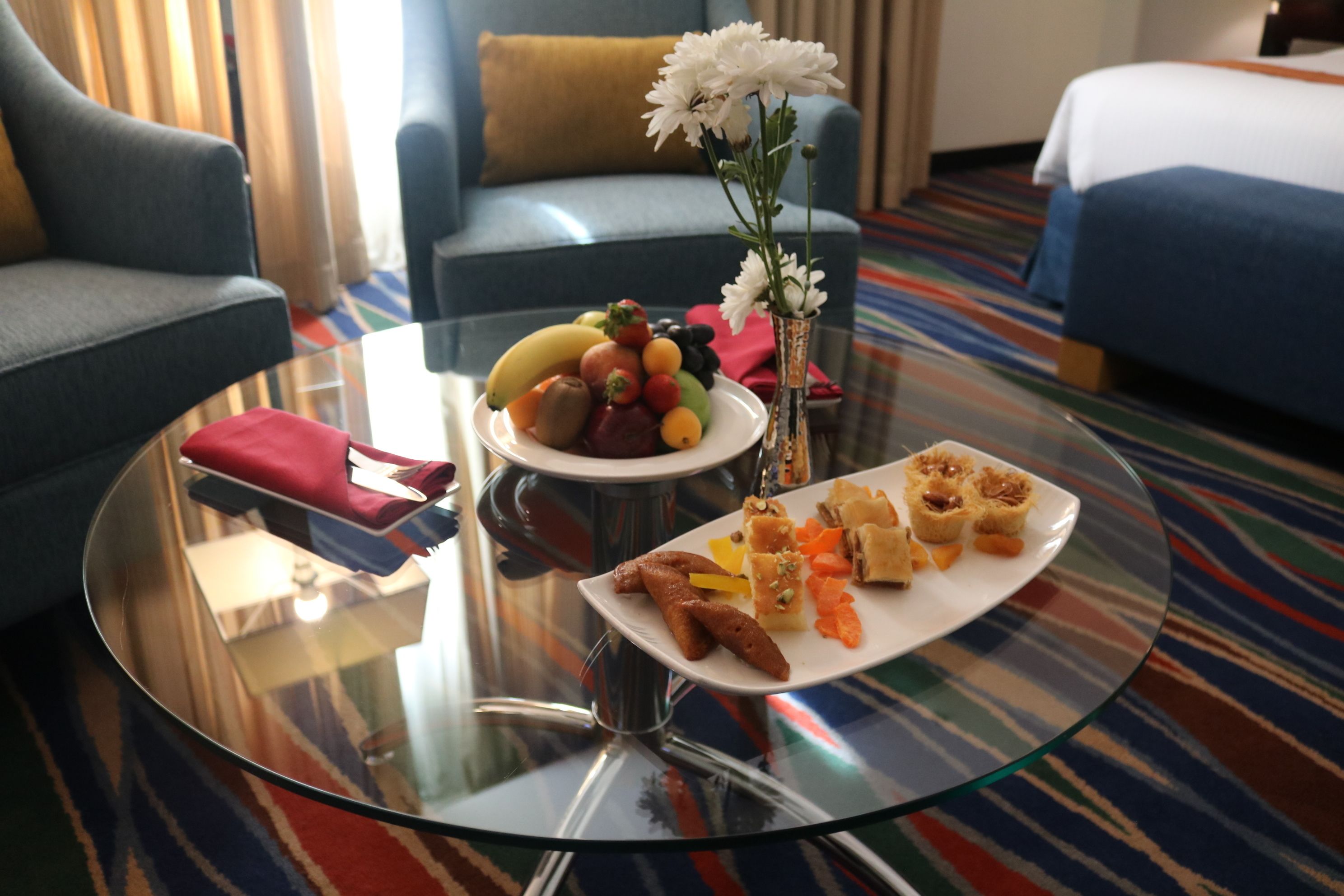 Habitació d'hotel amb taula de dinar de vidre que mostra un plat de fruita, un plat de pastissos i un ram de flors blanques.
