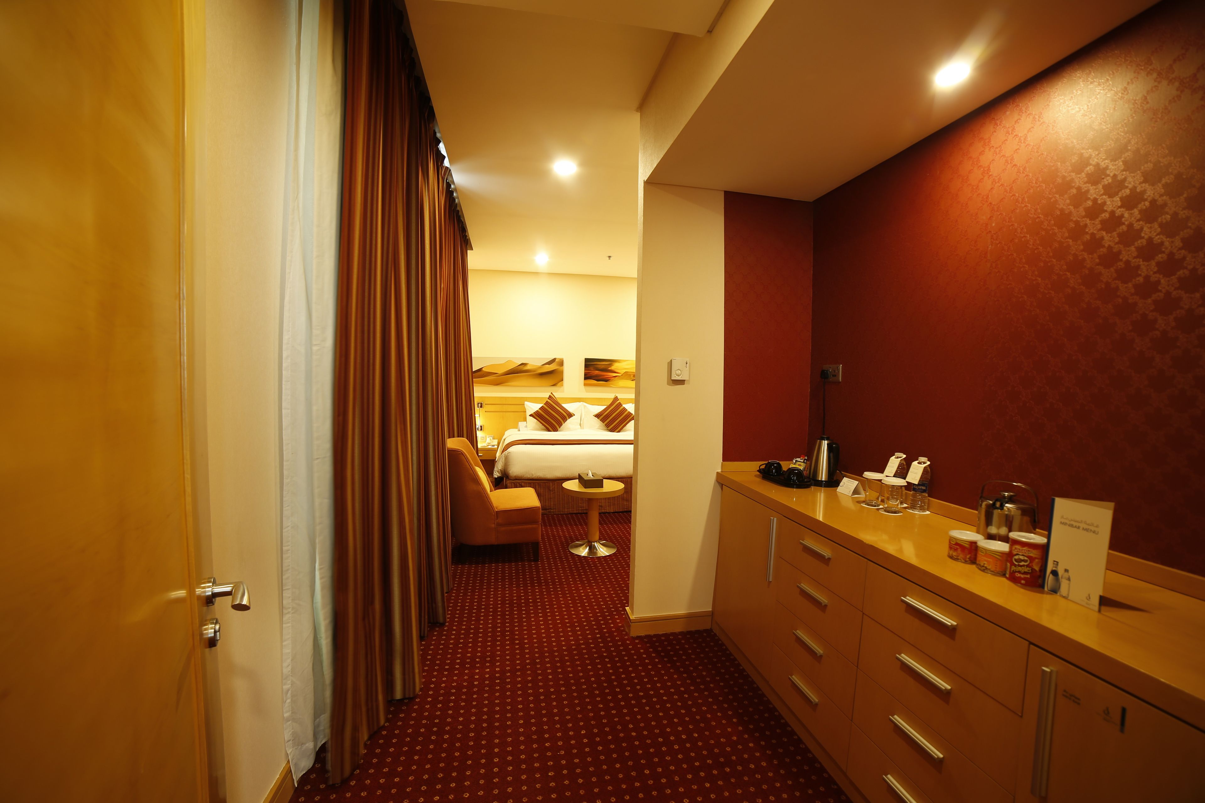 Habitación de hotel con cama, una mesita, una silla, un minibar y un baño con espejo y lavamanos.