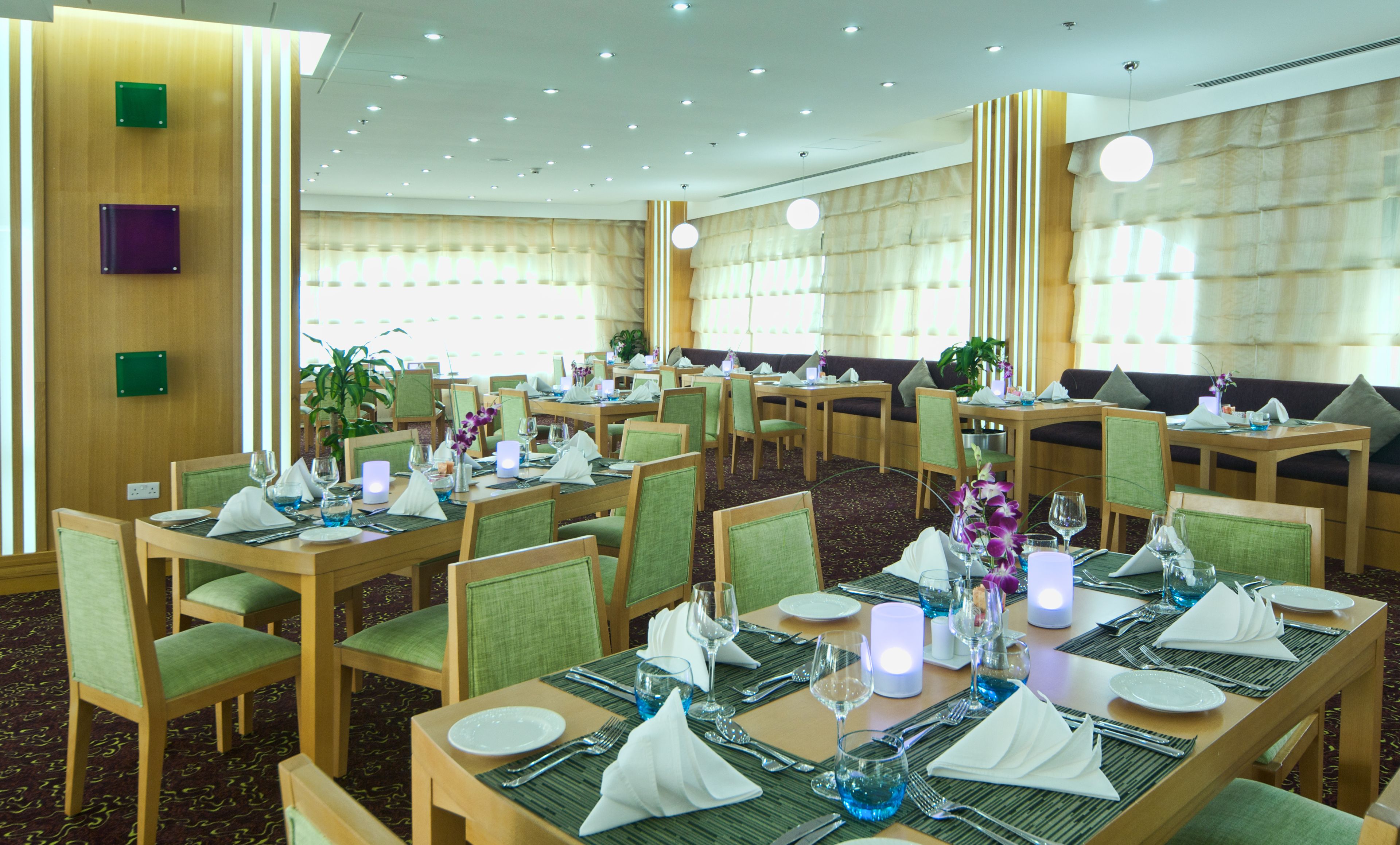 A hotel dining area with multiple tables set for guests, featuring green upholstered chairs and a carpeted floor.