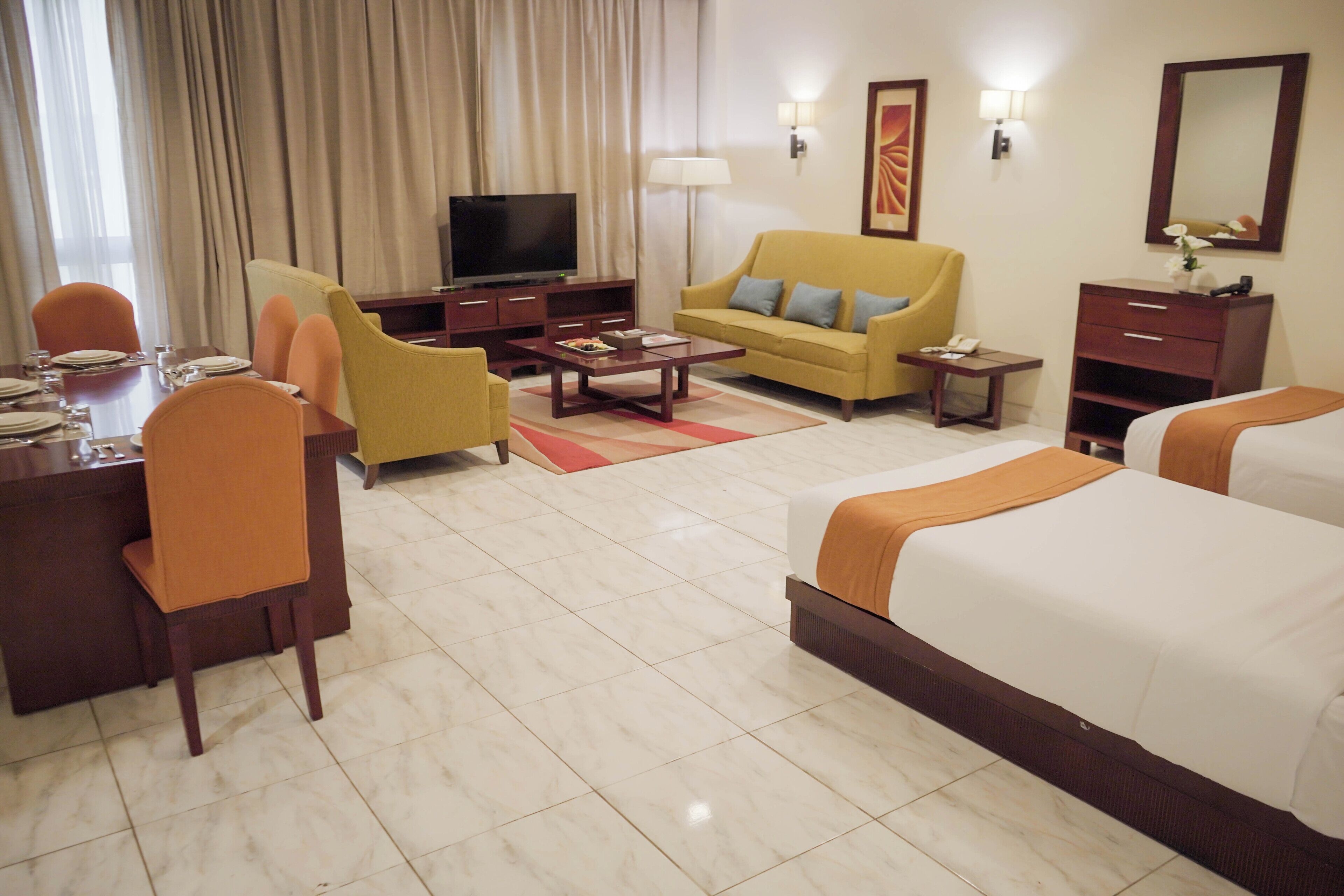 A hotel room with a bed, a sofa, a desk with a TV, and a dining area with chairs and a table.