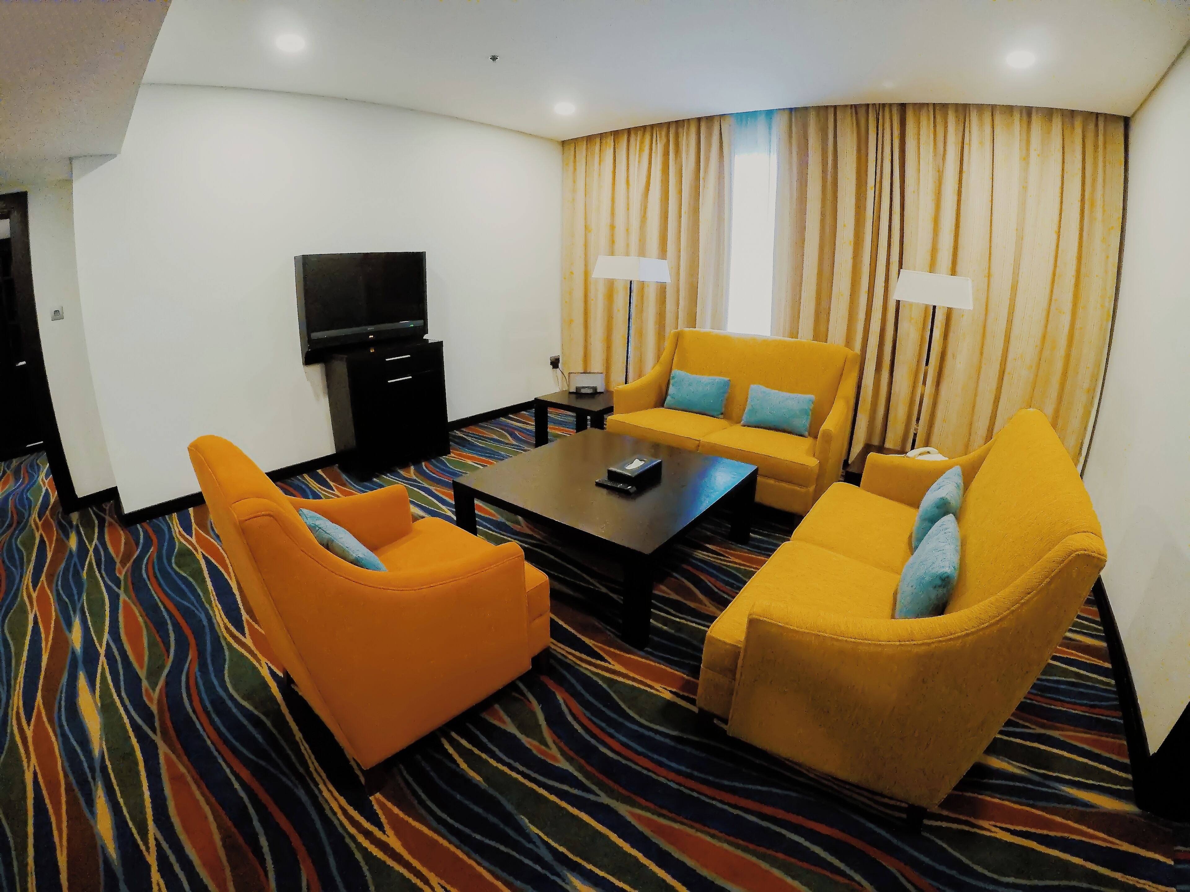 A hotel room with a sofa set, a coffee table, a flat-screen TV, and a patterned carpet.