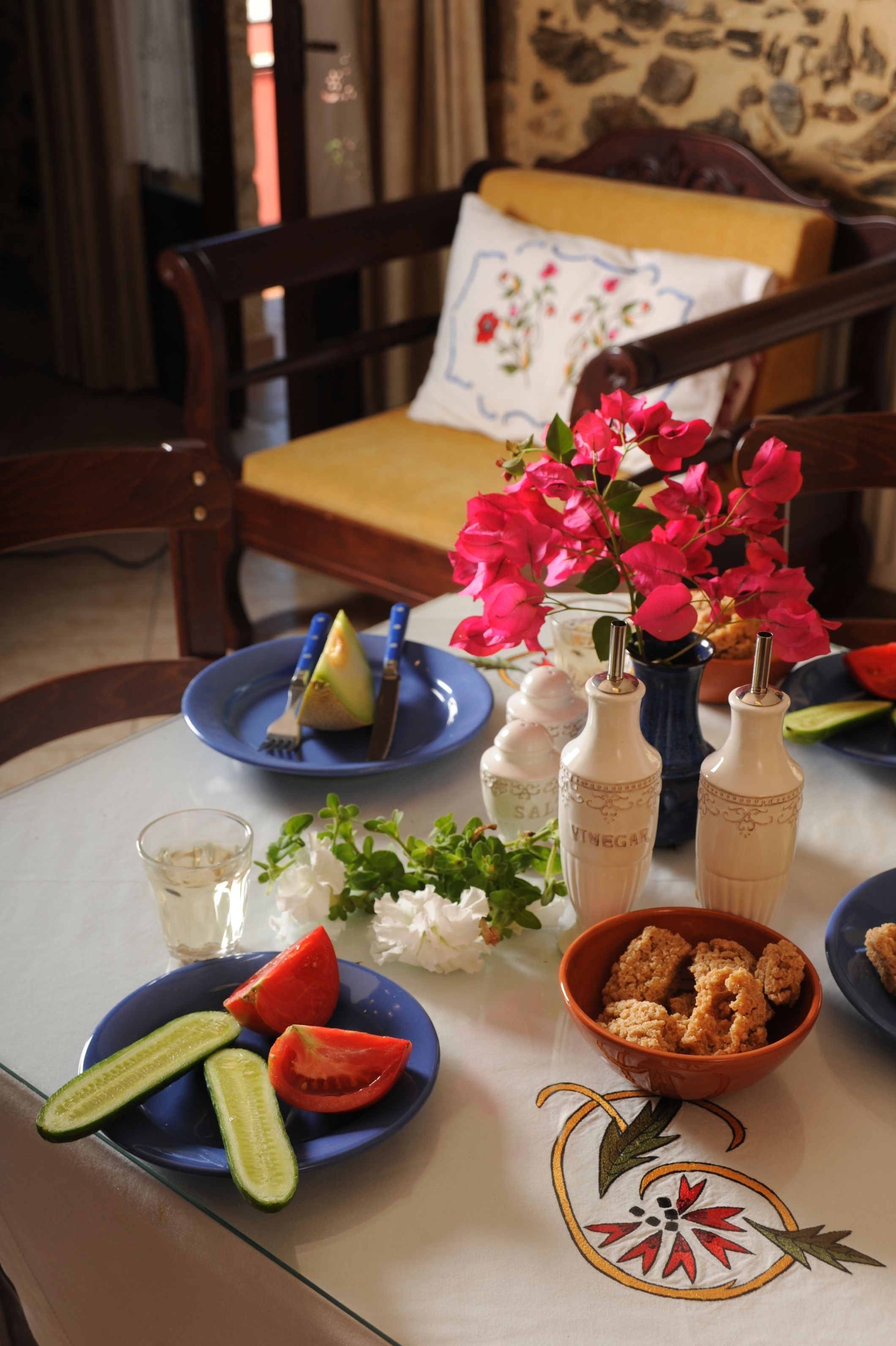 Una mesa de comedor con platos de comida, incluyendo rodajas de pepino y trozos de tomate, y un bol de croutons. También hay dos botellas con la etiqueta 'VINEGAR' y un florero con flores rosas.