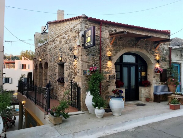 A traditional stone building with a tiled roof, a black door, and a sign reading "Jani Traditional House." There are potted plants and a bench outside. - Idili Traditional House (Mylopotamos)