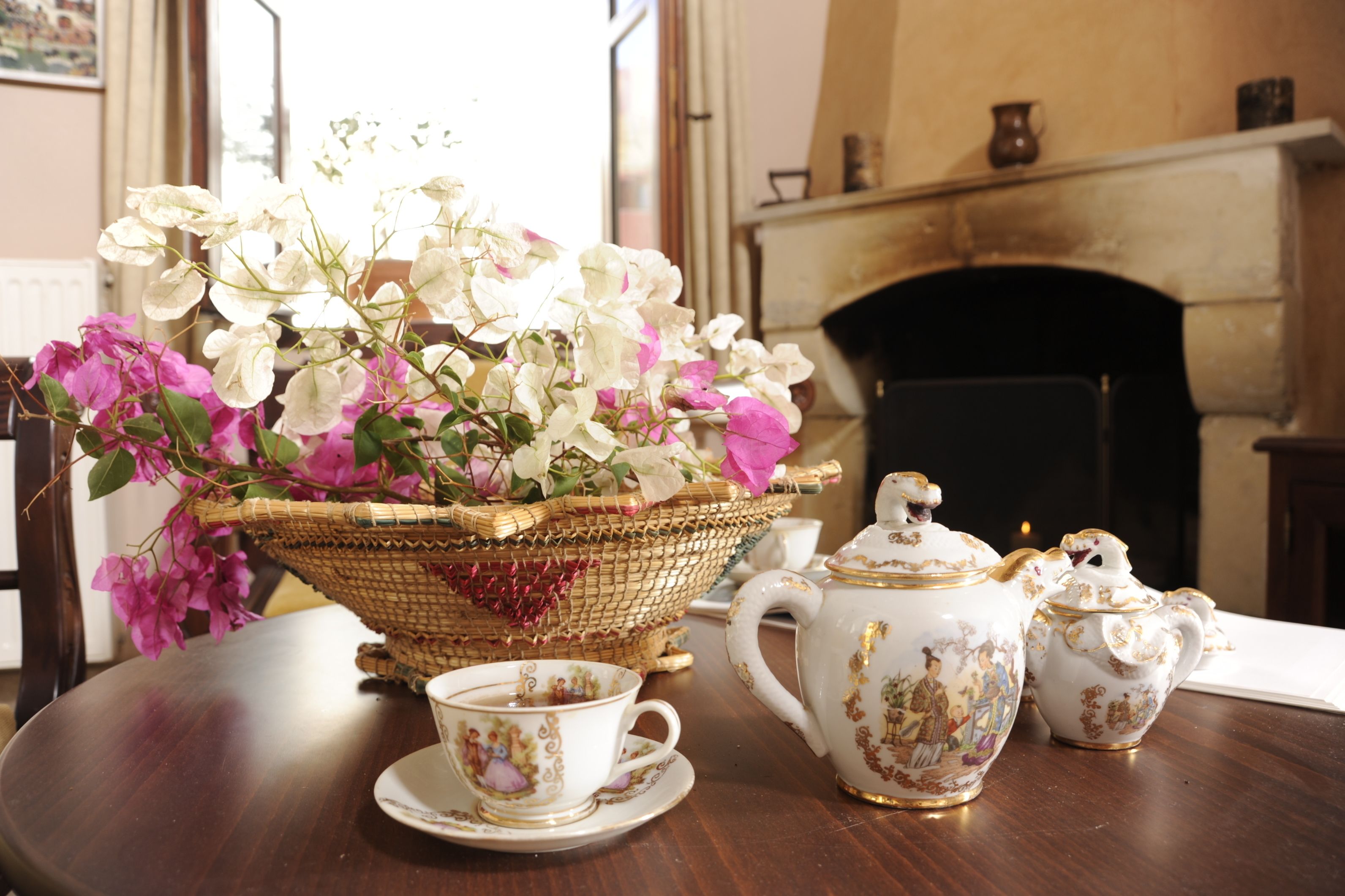 Une table sur laquelle se trouve un panier en osier contenant des fleurs blanches et roses, une théière et deux tasses avec leurs soucoupes.