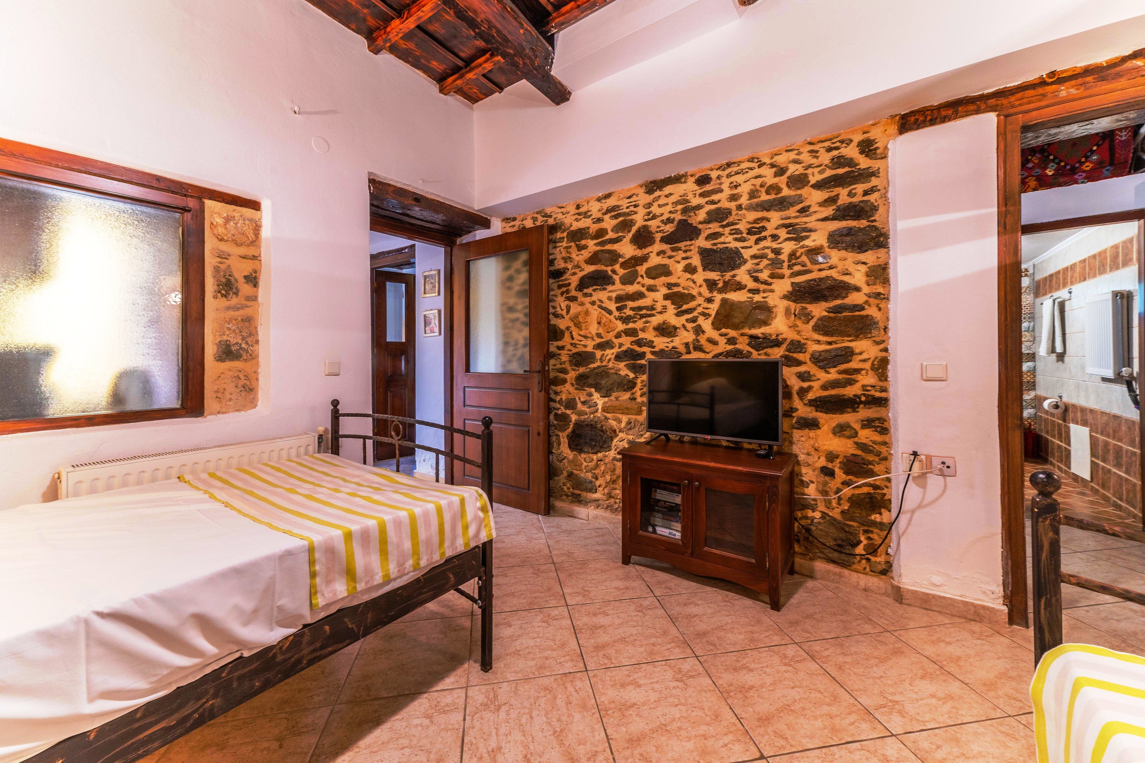 A bedroom with a bed, a television on a wooden stand, a stone wall, and a tiled floor.