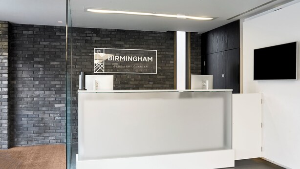 A reception area with a glass partition, a reception desk, a black brick wall, and a mounted television. - Four Points Flex by Sheraton Birmingham Jewellery Quarter (Birmingham)