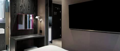 A modern hotel room with a large bed, a TV mounted on the wall, a mirror, and a small desk with a lamp.