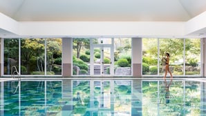 A large indoor swimming pool with clear glass walls, surrounded by a lush garden and a view of trees and rocks. - Aspria Royal La Rasante (Brussels)
