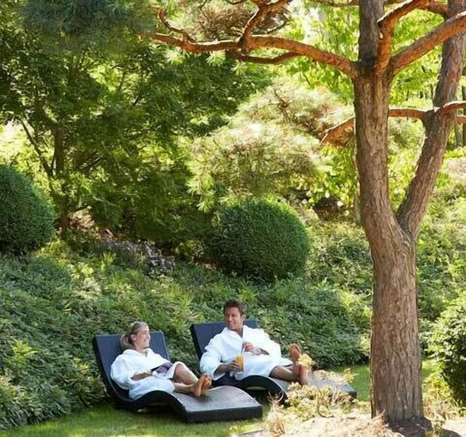 Two people relaxing on lounge chairs in a garden with dense greenery and a tree.