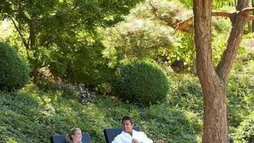 Two people relaxing on lounge chairs in a garden with dense greenery and a tree.