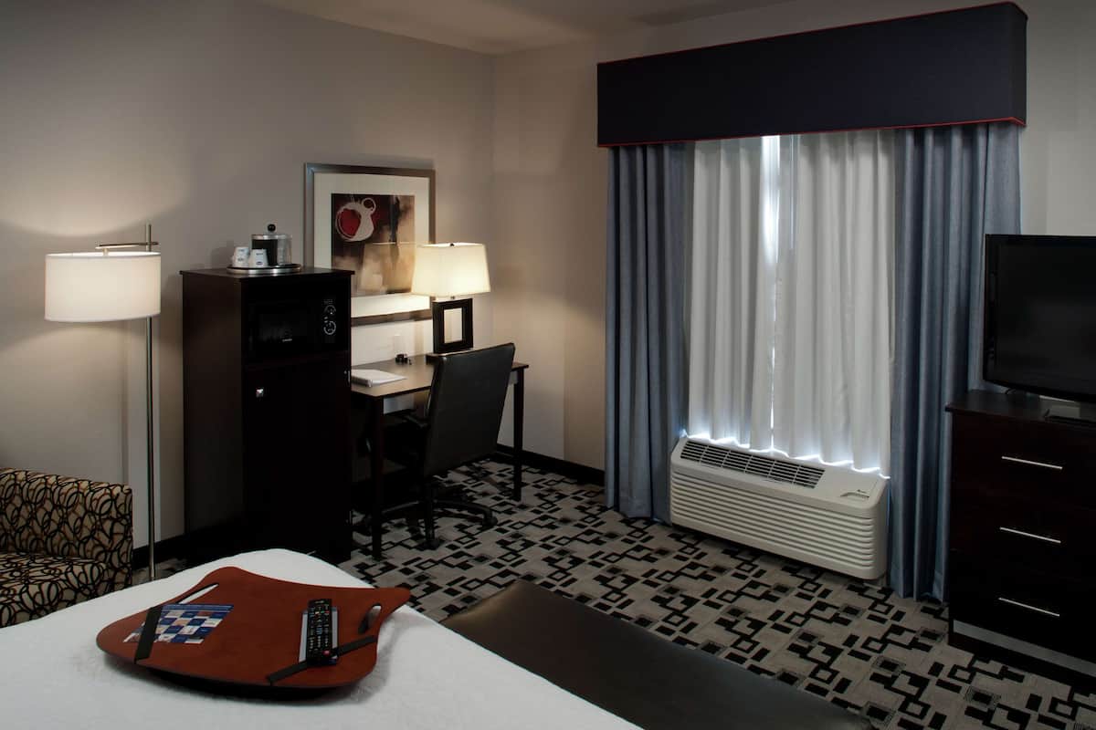A hotel room with a bed, desk, chair, lamp, television, and a patterned floor.