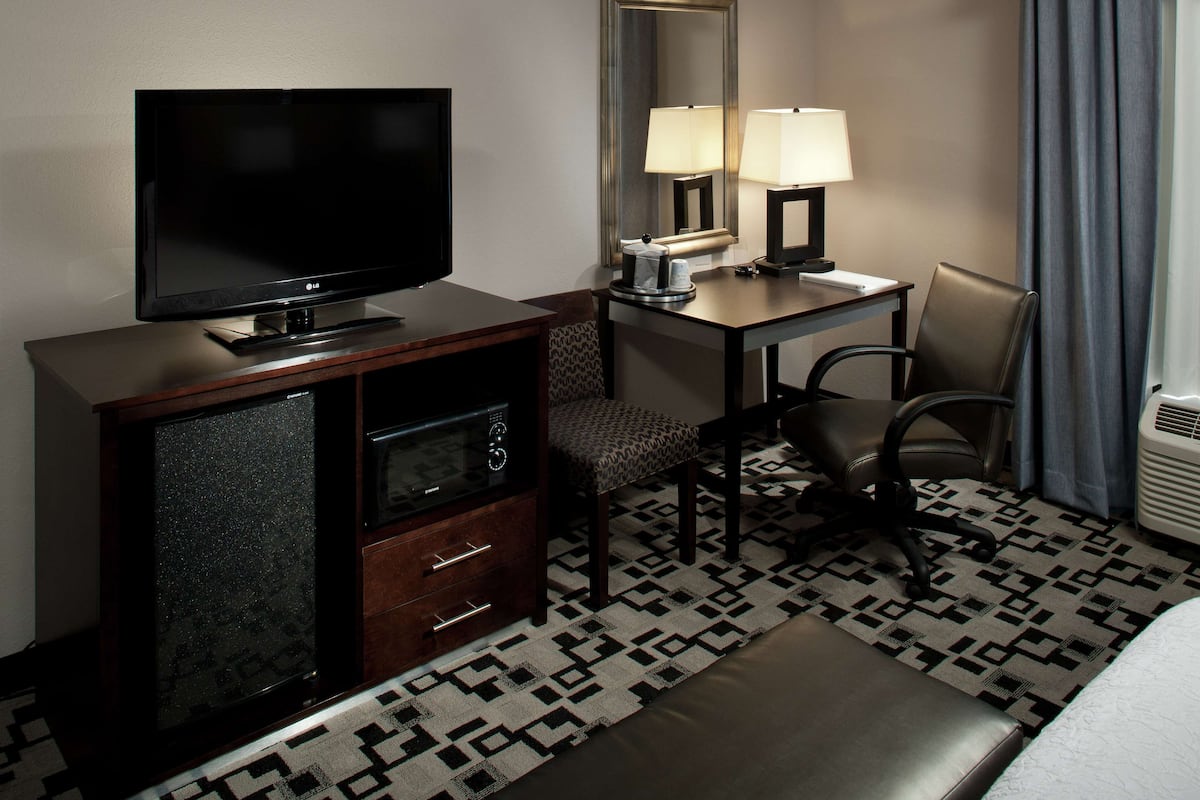 A hotel room with a flat-screen TV, a desk with a lamp, a chair, a sofa, and a patterned rug.
