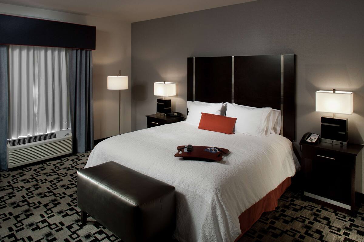 A hotel room with a large bed, two bedside tables with lamps, a small desk, a chair, and a window with curtains.
