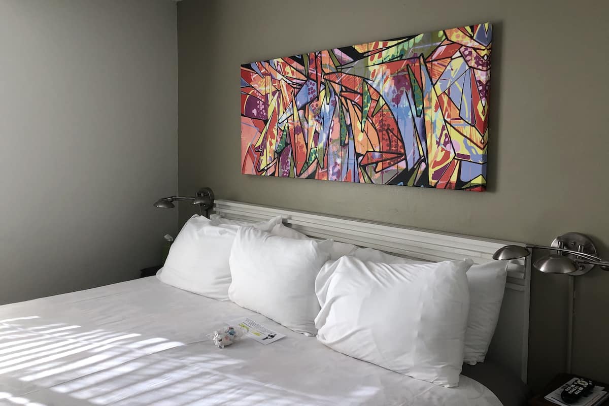 Habitación de hotel con un cuadro abstracto grande en la pared, una cama bien hecha con ropa blanca y dos lámparas de noche.