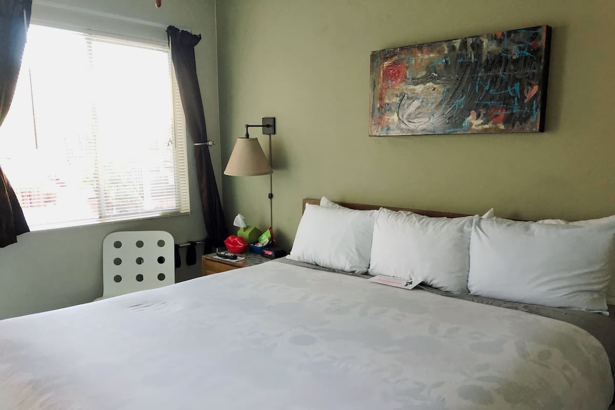 Habitación de hotel con una cama grande, una mesita de noche con lámpara, un cuadro en la pared y una ventana con cortinas.