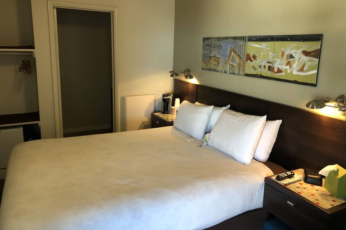 Una habitación de hotel con una cama grande, una mesita de noche con lámpara, un cuadro en la pared y una puerta a la izquierda.