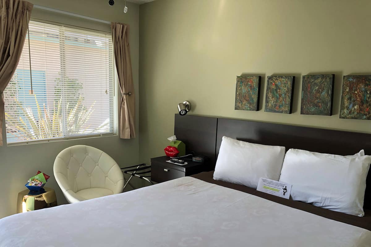 Habitación de hotel con una cama grande, una silla, una mesita de noche con lámpara y una ventana con cortinas. Hay tres cuadros en la pared sobre la cama.