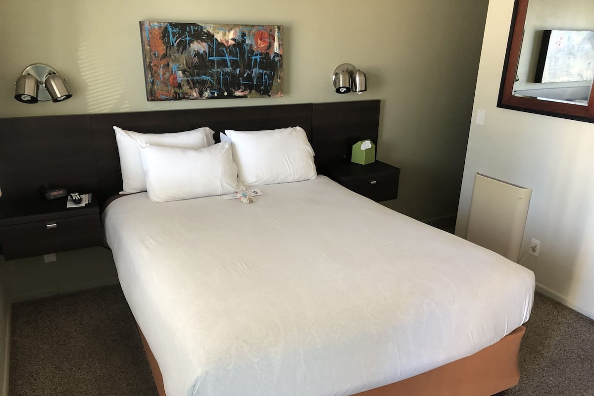 Una cama bien hecha con ropa de cama blanca, un cabecero oscuro y un cuadro en la pared. Hay dos mesitas de noche con lámparas y una caja verde en una de ellas.
