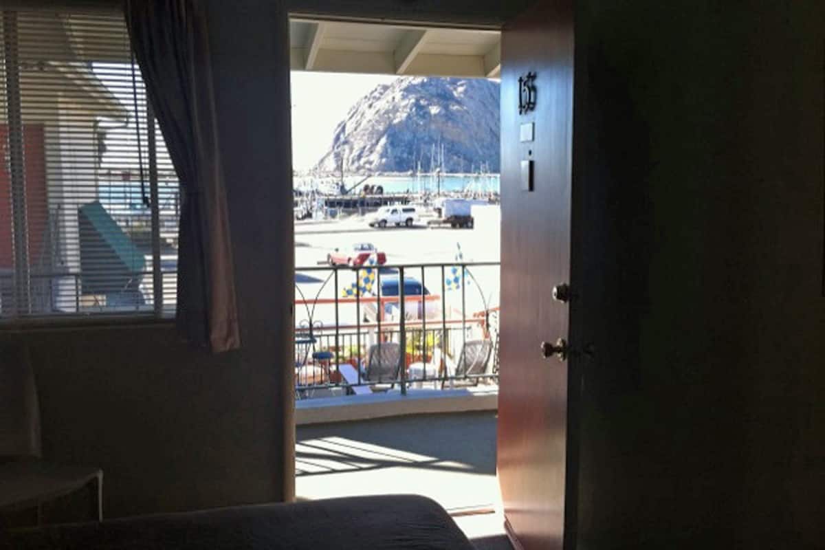 Una habitación con una cama, un ventanal con persianas y vista a un estacionamiento con coches y una montaña al fondo.
