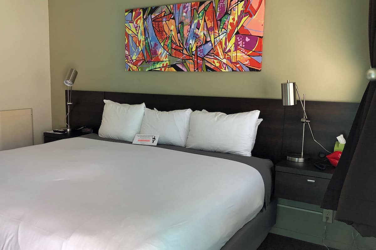Habitación de hotel con una cama grande, ropa de cama blanca, una mesita de noche con una lámpara, una mesita con una caja de pañuelos y un cuadro abstracto y colorido en la pared.