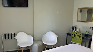 A hotel room with a bed, two white chairs, a small table with a green chair, a radiator, a mirror, and a TV on the wall.