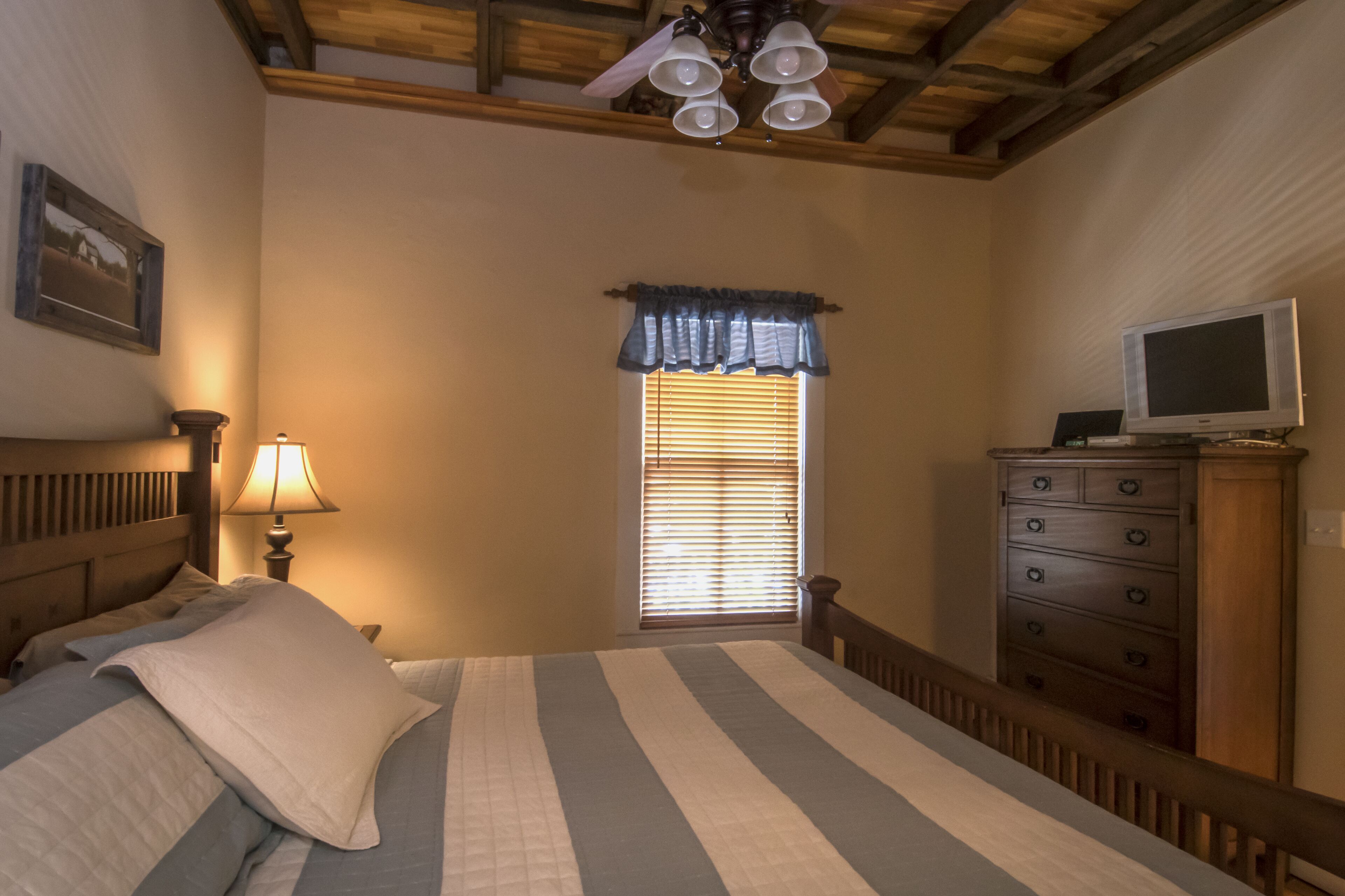 Un dormitorio con cama, cómoda, televisor, ventana con persianas y ventilador de techo.