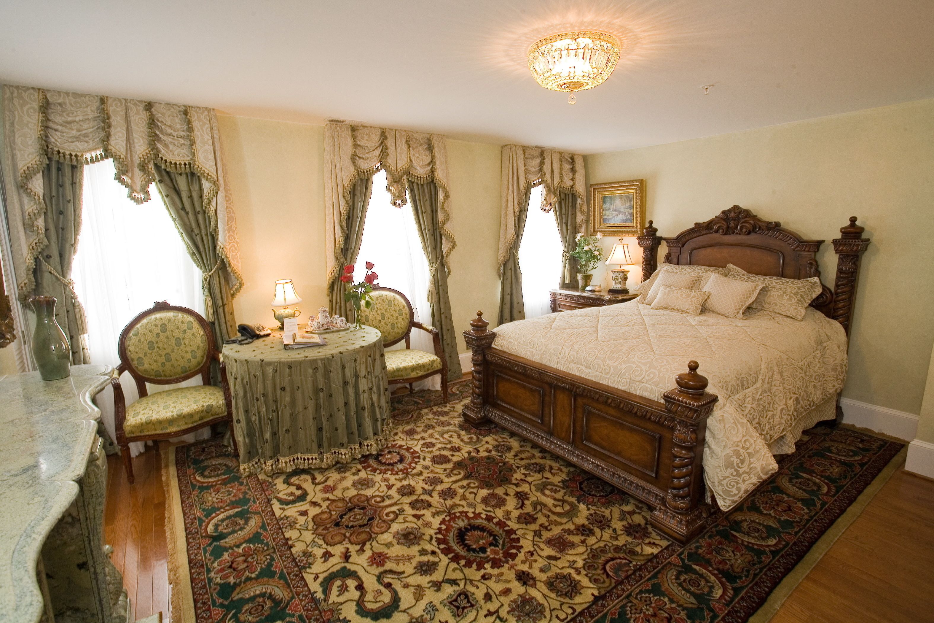 A bedroom with a large bed, a round table, two chairs, a chandelier, and a patterned carpet.