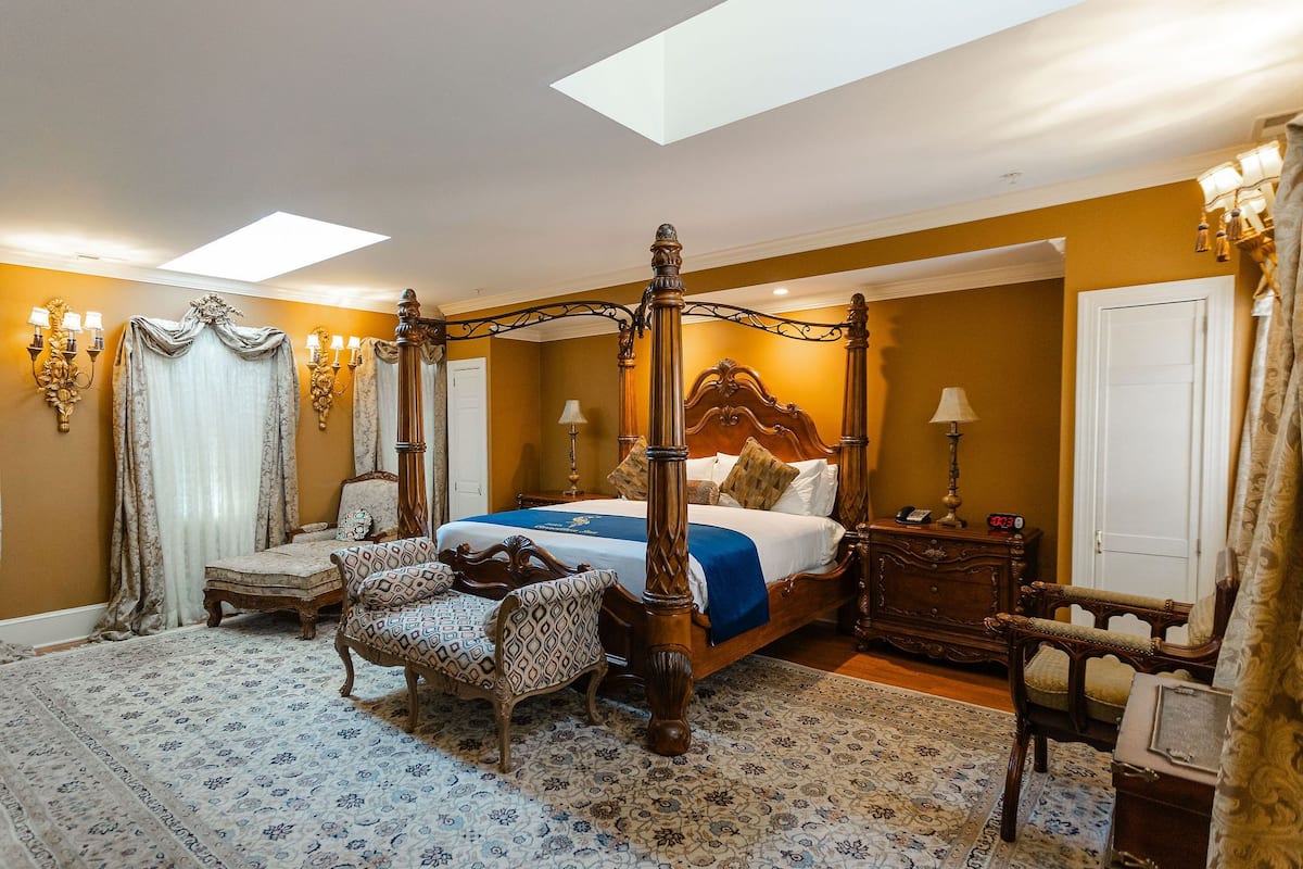 A four-poster bed with a canopy, a sitting area with patterned chairs, a bedside table with a lamp, and a large window with natural light.