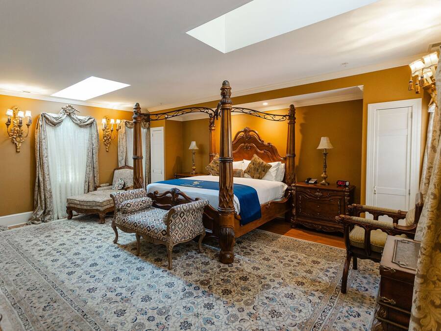 A four-poster bed with a canopy, a sitting area with patterned chairs, a bedside table with a lamp, and a large window with natural light.