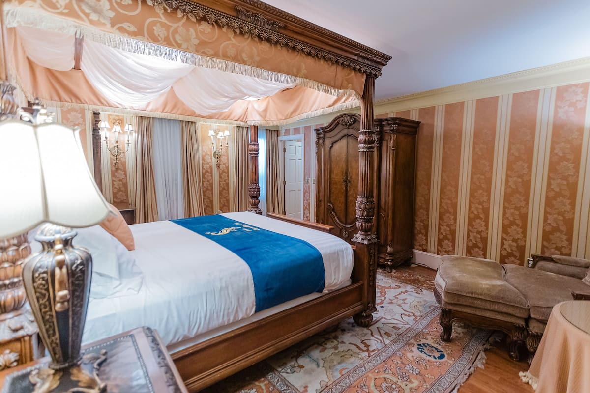 A classic four-poster bed with a blue accent, a wooden nightstand, a vintage lamp, and a patterned rug.