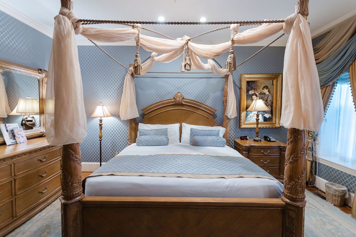 A four-poster canopy bed with a wooden headboard, a nightstand with a lamp, and a mirror on the dresser.