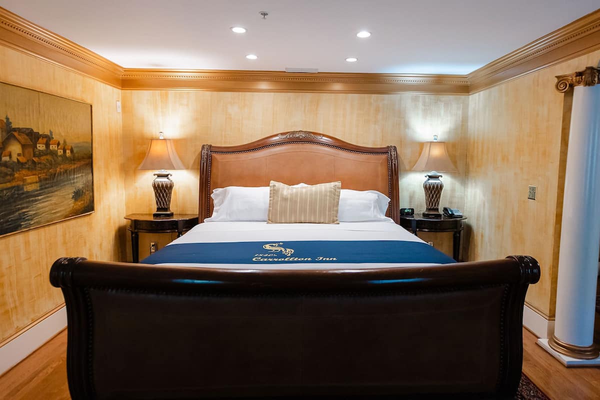 A large bed with a wooden headboard, two bedside tables with lamps, a painting on the wall, and a carpeted floor.