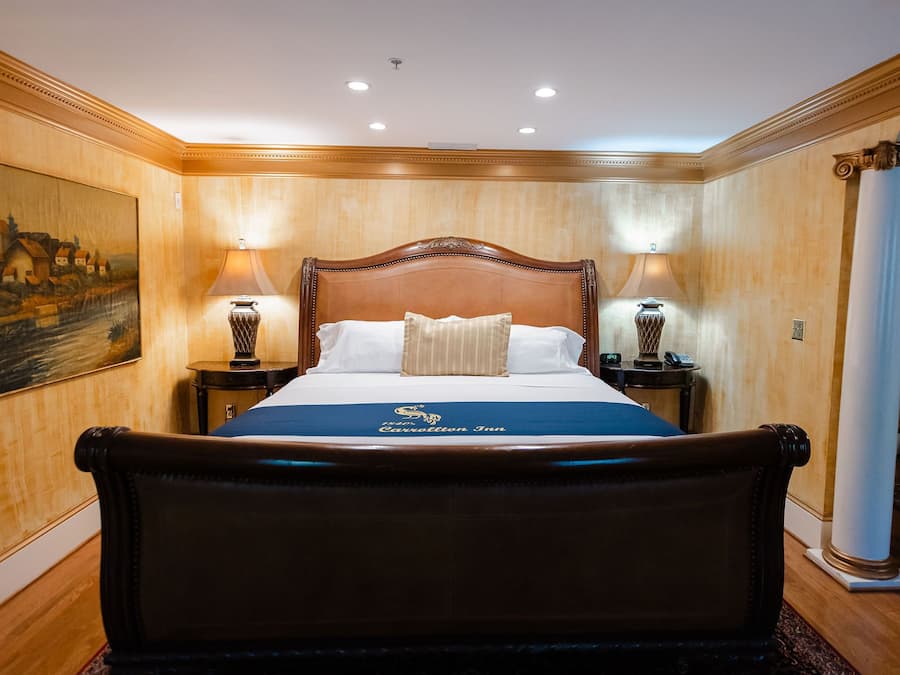 A large bed with a wooden headboard, two bedside tables with lamps, a painting on the wall, and a carpeted floor.
